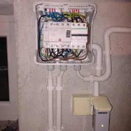 Otwarta skrzynka elektryczna z bezpiecznikami Eaton i Schneider, widoczne przewody, rury PCV i urządzenie Correx z zieloną diodą, zamontowana na szarej ścianie w pobliżu okna.