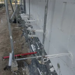 Montaż przewodów w ścianie z ociepleniem, widoczne rusztowanie, poziomica i pistolet do pianki montażowej.