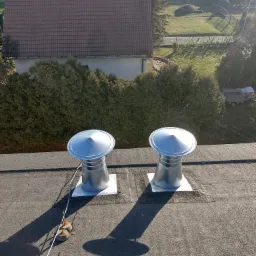 Dwa nowe, metalowe kominki wentylacyjne z daszkami na płaskim dachu pokrytym papą, zabezpieczone linką z obciążnikami, z widokiem na dom i zielone otoczenie.