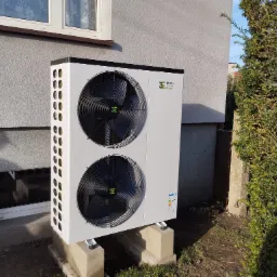 Biała, dwuwentylatorowa jednostka zewnętrzna z logo na ścianie budynku, zamontowana na betonowych podstawach, obok krzewu.