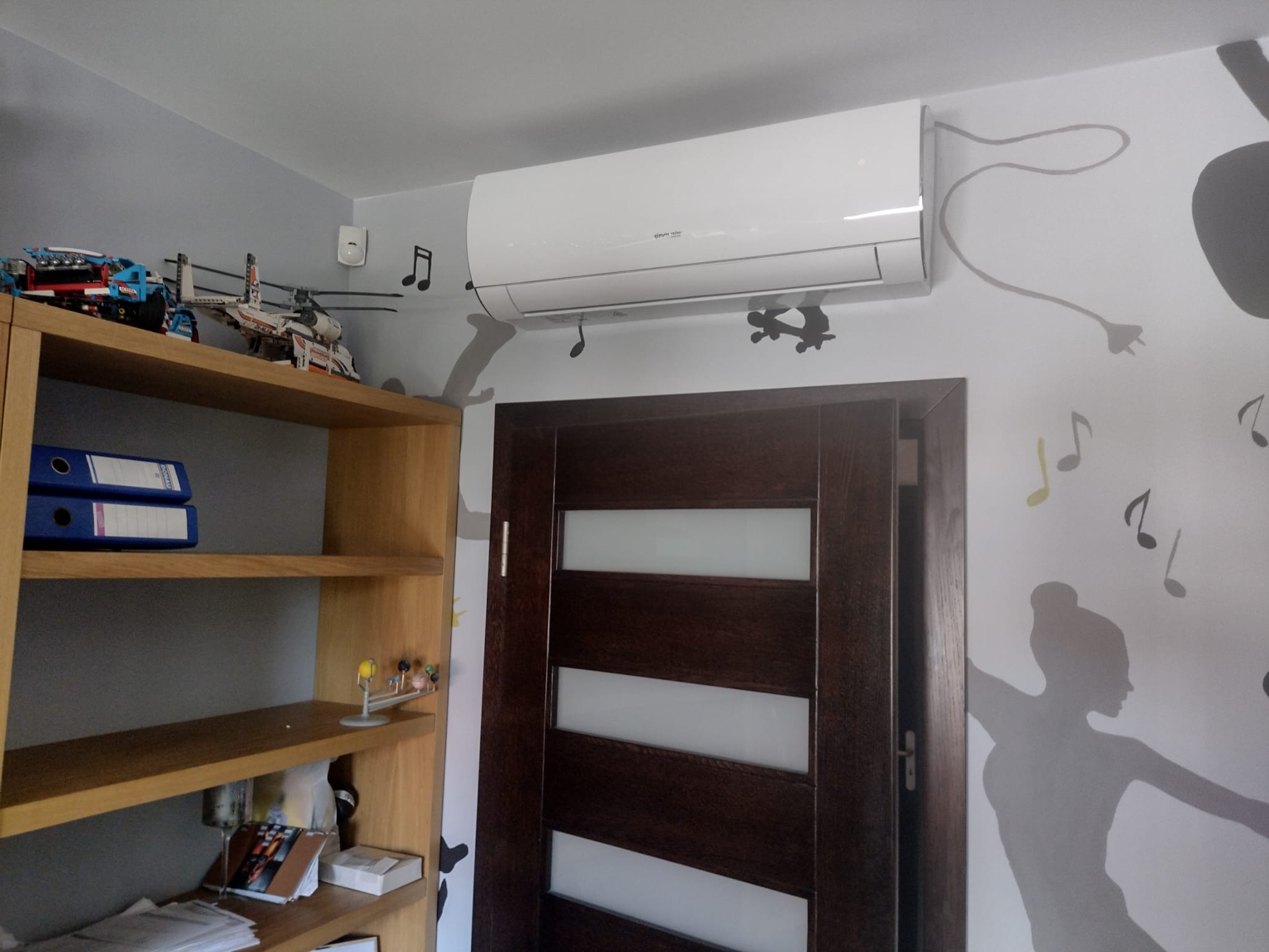 Biały klimatyzator ścienny zamontowany nad ciemnymi drzwiami w pokoju z półkami wypełnionymi książkami i zabawkami, ściana ozdobiona naklejkami z motywem muzycznym.