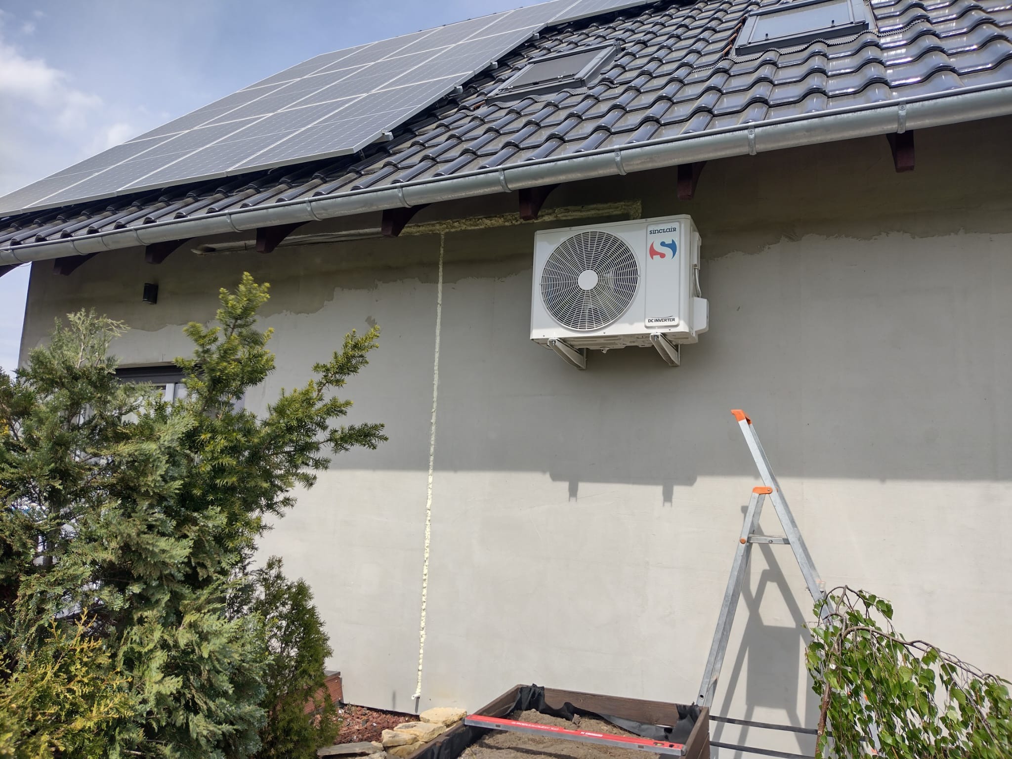 Zewnętrzna jednostka klimatyzatora Sinclair zamontowana na ścianie domu z panelami słonecznymi na dachu, widoczna drabina i ślady pianki montażowej.