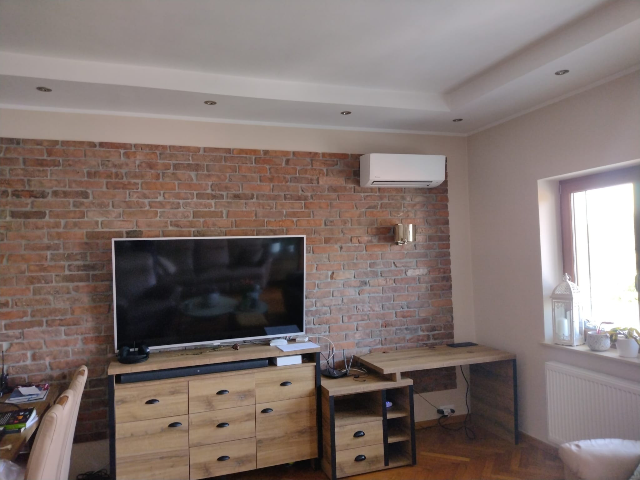 Biały klimatyzator ścienny zamontowany na ceglanej ścianie w salonie, widok na szafkę RTV, biurko i okno z dekoracjami.