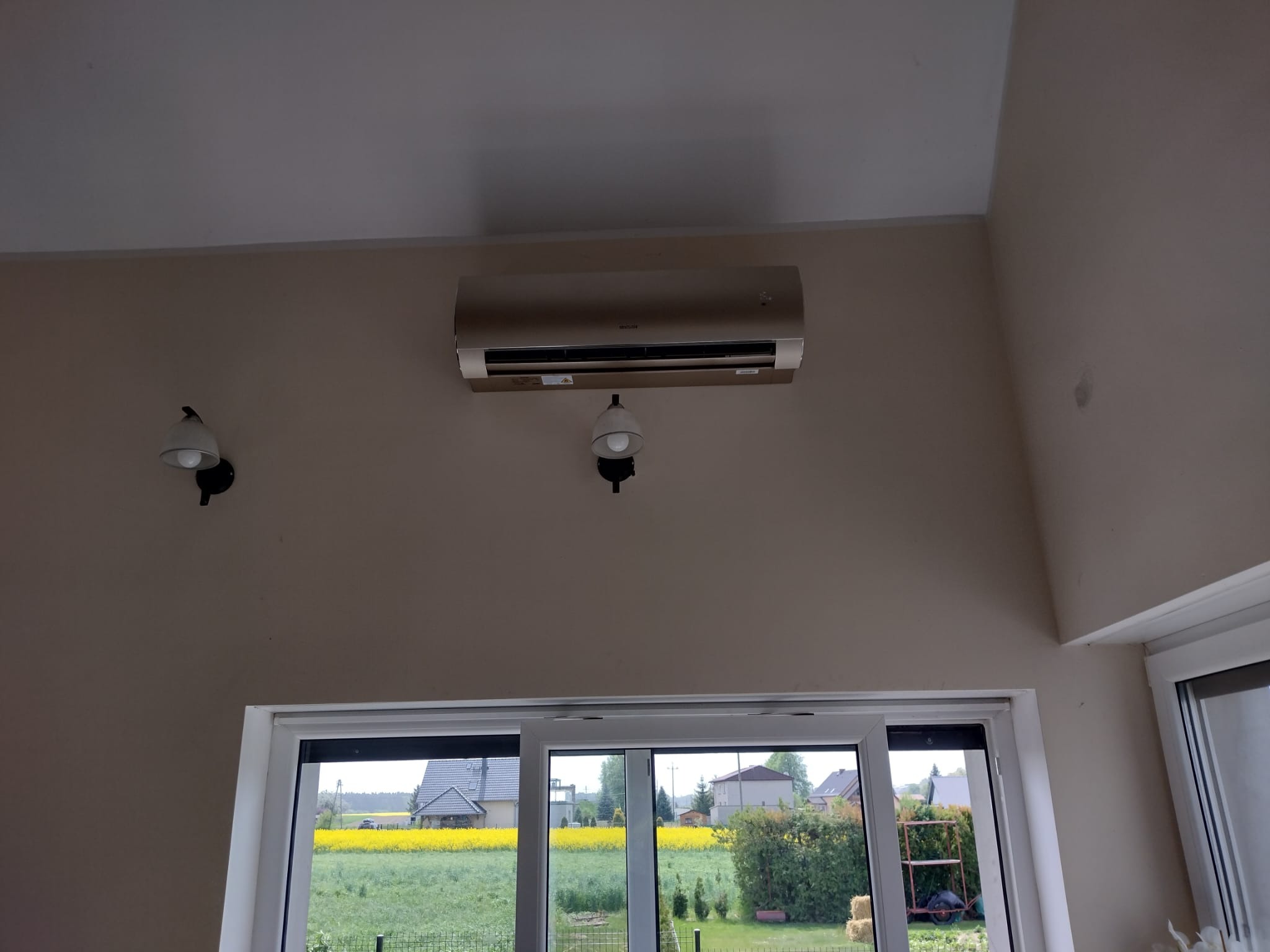 Zamontowany klimatyzator ścienny w jasnym wnętrzu domu, widok przez okno na kwitnące pole rzepaku.