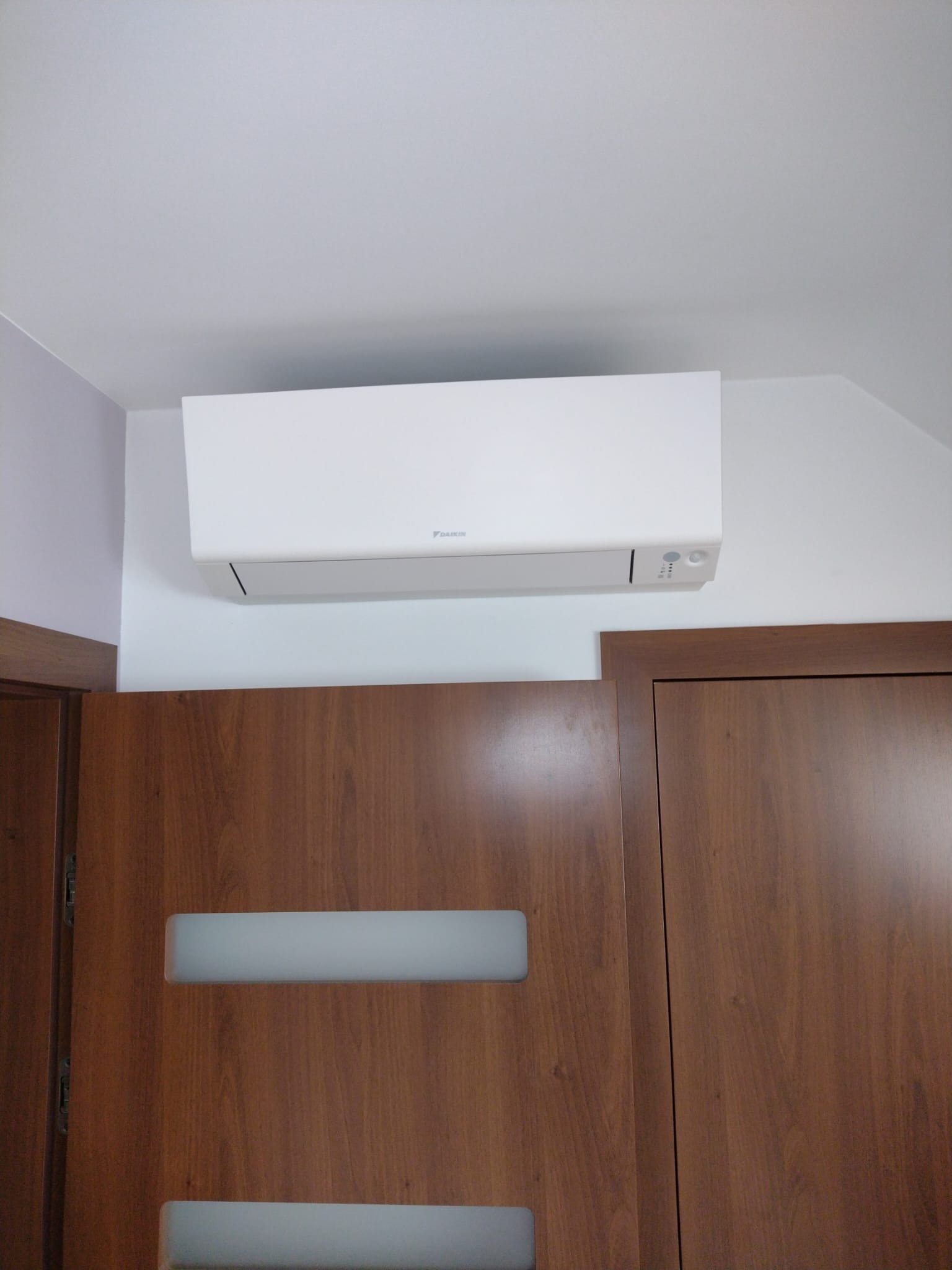Biały klimatyzator Daikin zamontowany na ścianie nad drewnianymi drzwiami z mlecznymi szybami, widok z dołu.