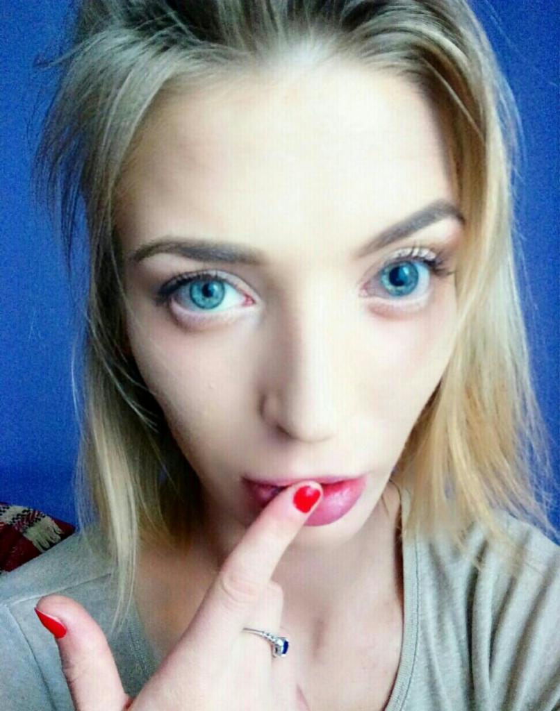 Portret kobiety z blond włosami i niebieskimi oczami, z czerwonym manicurem i pierścionkiem na palcu, na tle niebieskiej ściany. Usta pomalowane czerwoną szminką.