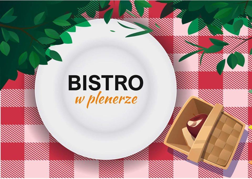 Grafika przedstawiająca biały talerz z napisem 'BISTRO w plenerze', koszyk piknikowy z jabłkiem, całość na czerwono-białym obrusie w kratę, z dekoracjami z zielonych gałązek.