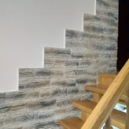 Ściana przy schodach wykończona dekoracyjną okładziną imitującą kamień, częściowo przylegająca do gładkiej, białej ściany, widoczne drewniane schody.
