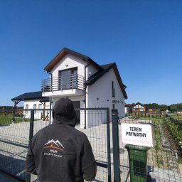 Paweł Wójcik W&W - Nowoczesny dom jednorodzinny z balkonem, widziany zza ogrodzenia przez osobę w bluzie z logo firmy budowlanej na tle błękitnego nieba. Tabliczka 'Teren prywatny'.