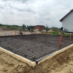 Paweł Wójcik W&W - Zbrojenie fundamentu domu jednorodzinnego z widoczną izolacją termiczną i wystającymi rurami instalacyjnymi. W tle budynek w stanie surowym otwartym.