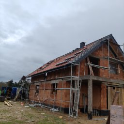 Paweł Wójcik W&W - Budowa domu z czerwonej cegły w trakcie krycia dachu, widoczne rusztowania i częściowo ułożona dachówka na tle pochmurnego nieba. Etap prac dekarskich.