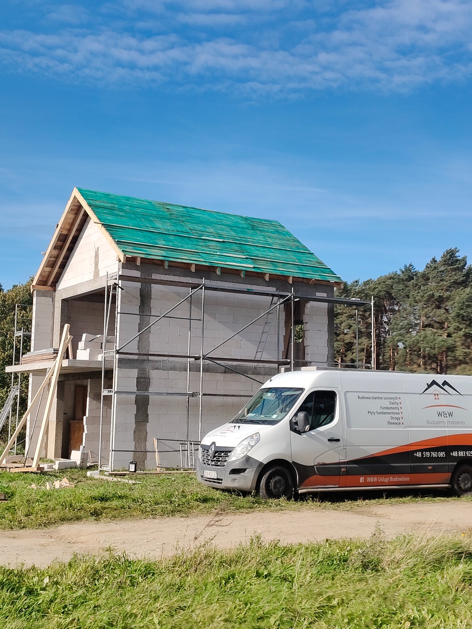 Budowa domu z białego bloczka, z zielonym dachem w trakcie krycia, na tle błękitnego nieba. Obok zaparkowany biały bus z logo firmy budowlanej.
