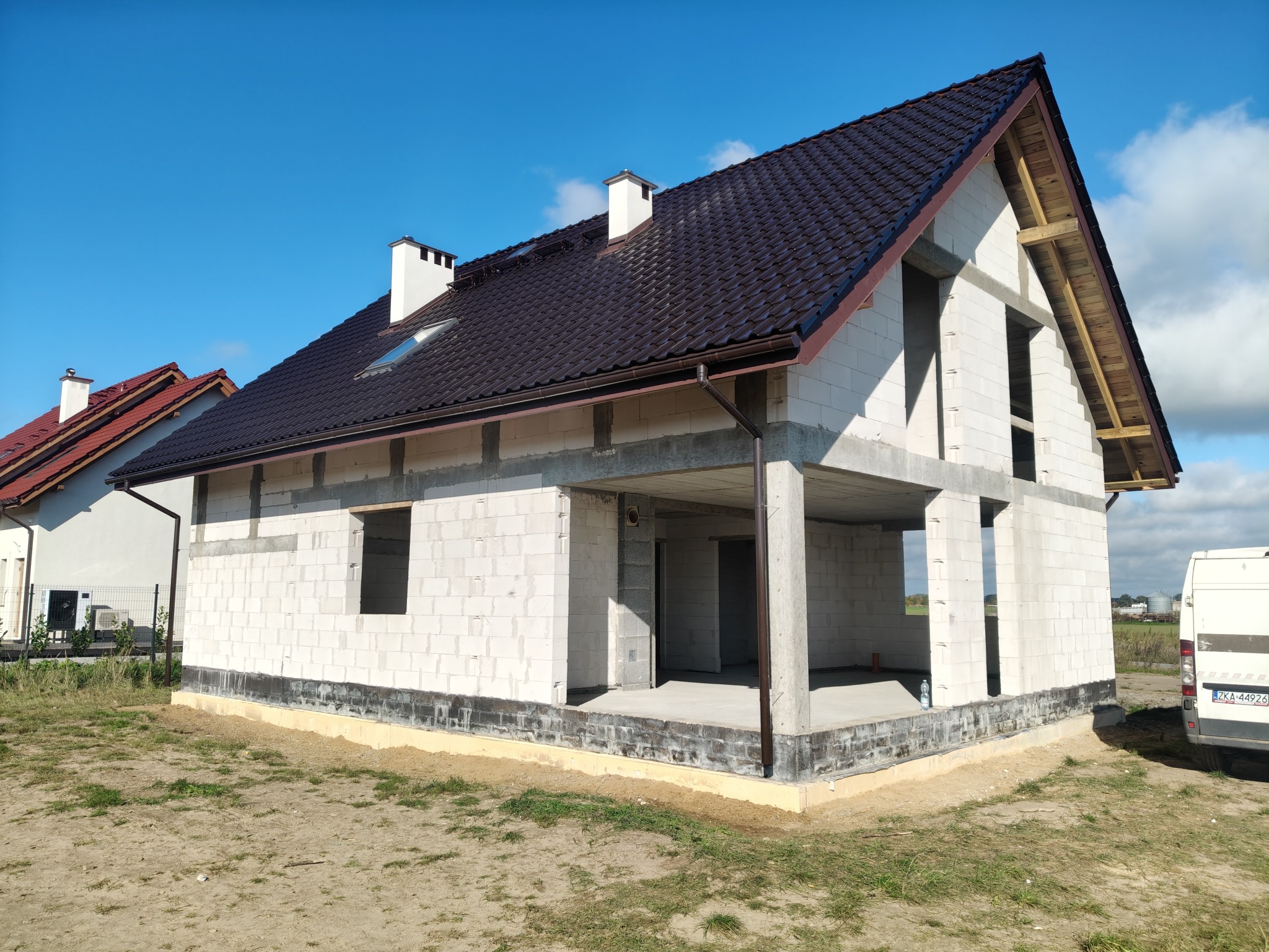 Surowy dom jednorodzinny w trakcie budowy, z ciemnym dachem i białymi ścianami z betonu komórkowego, na tle błękitnego nieba. Widoczne elementy konstrukcji dachu.