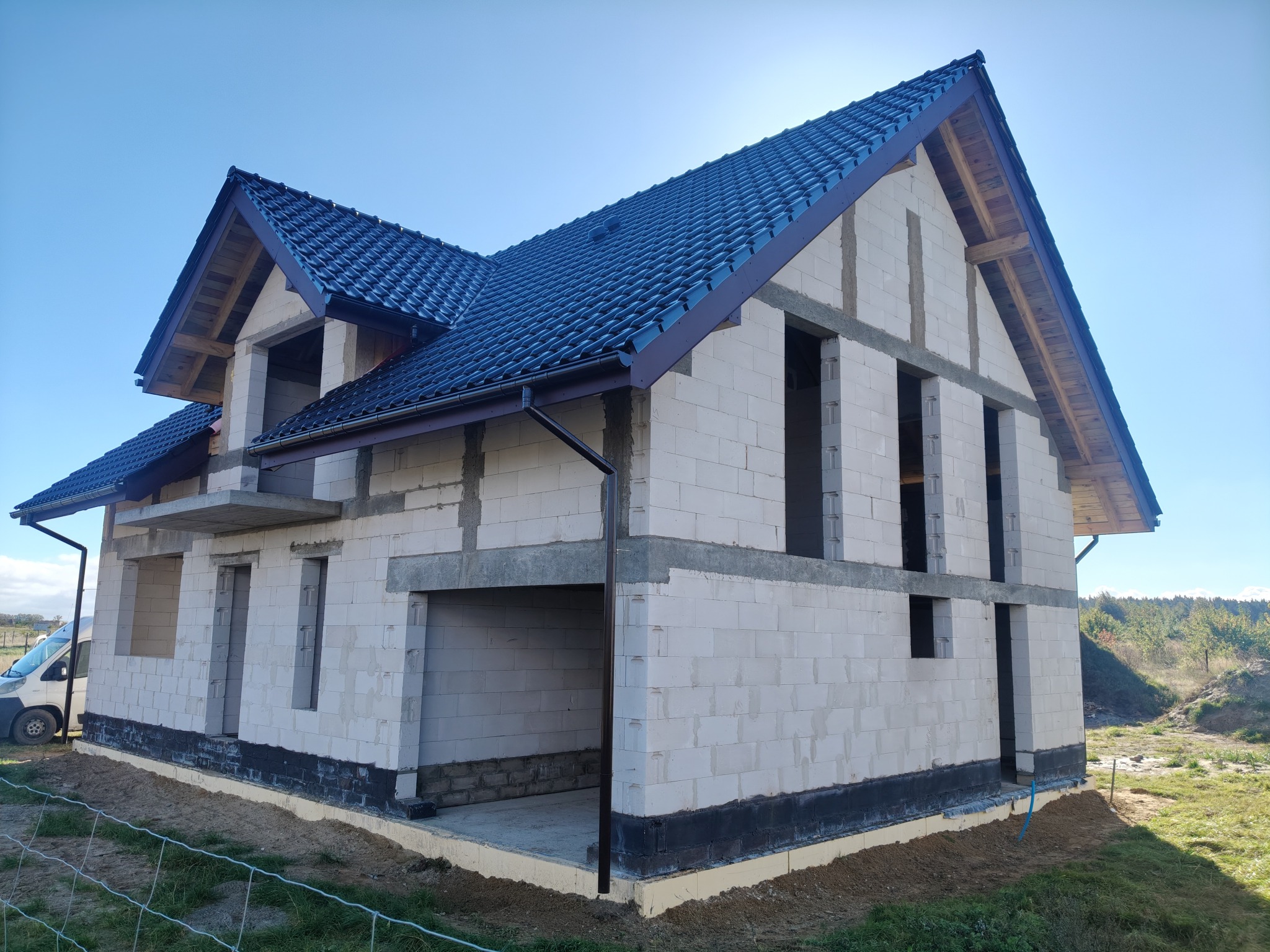 Surowy dom z białego betonu komórkowego pod ciemną dachówką, z widocznymi elementami konstrukcyjnymi i oknami bez szyb, na tle błękitnego nieba i zieleni.