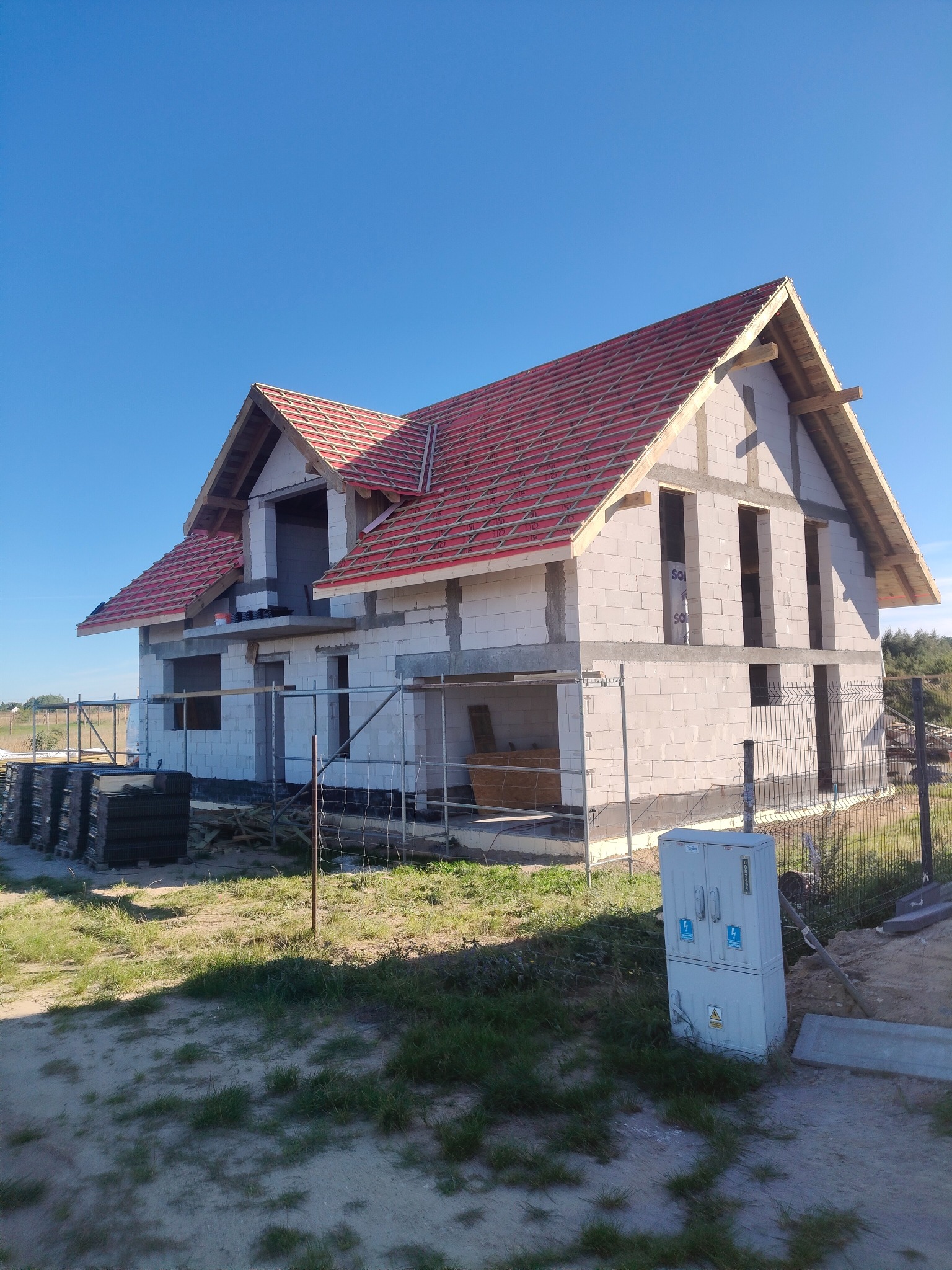 Surowy dom z białego bloczka w trakcie budowy, z częściowo ułożonym czerwonym dachem i widocznymi elementami konstrukcyjnymi. Błękitne niebo w tle.