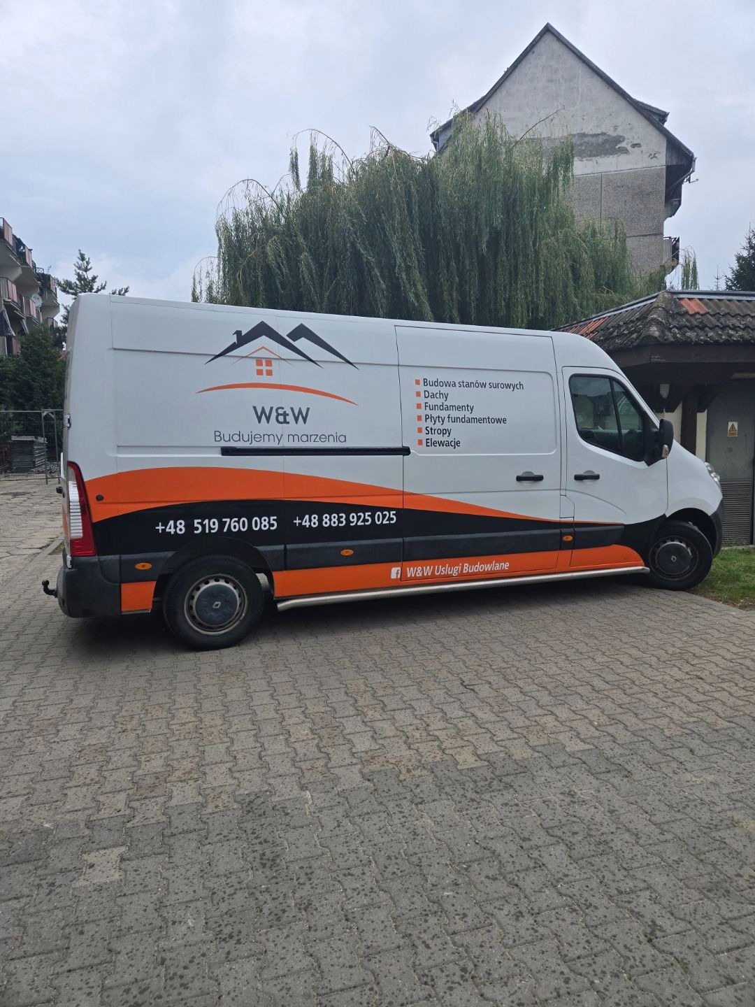Biały van z logo firmy budowlanej W&W, z pomarańczowymi i czarnymi akcentami, zaparkowany na brukowanym placu. W tle budynek i drzewo.