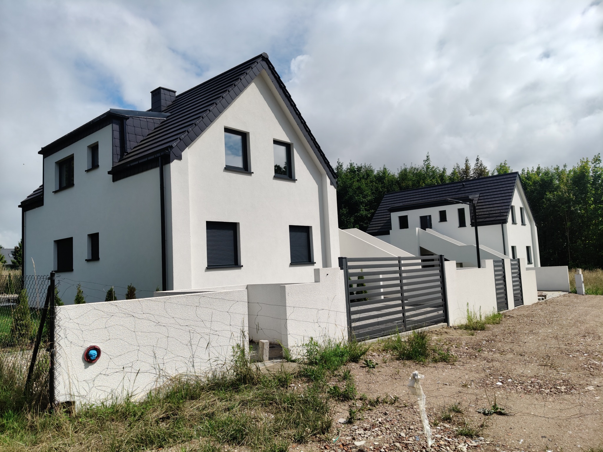 Nowoczesny dom jednorodzinny z czarnym dachem i białymi ścianami, otoczony niskim murem i metalowym ogrodzeniem. Widok na drugi dom w tle, pochmurne niebo.