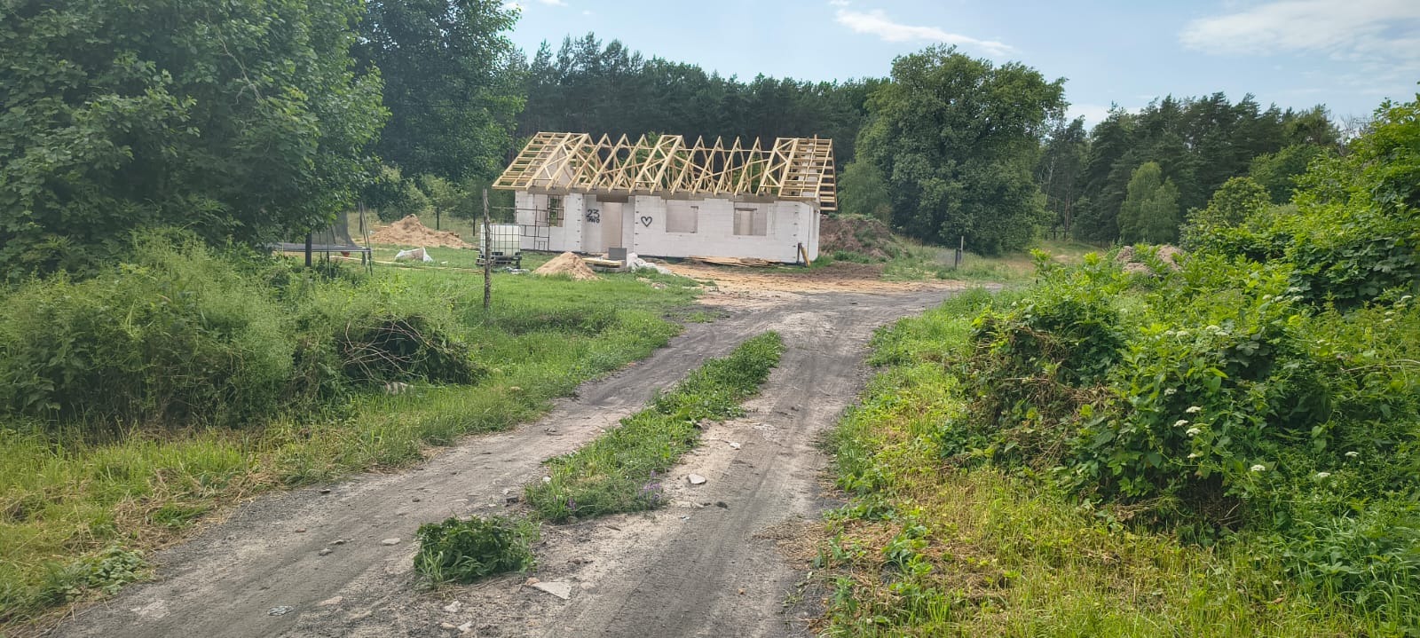Budowa domu jednorodzinnego w stanie surowym otwartym, widok z polnej drogi. Białe ściany z pustaków, drewniana konstrukcja dachu, zielone otoczenie.