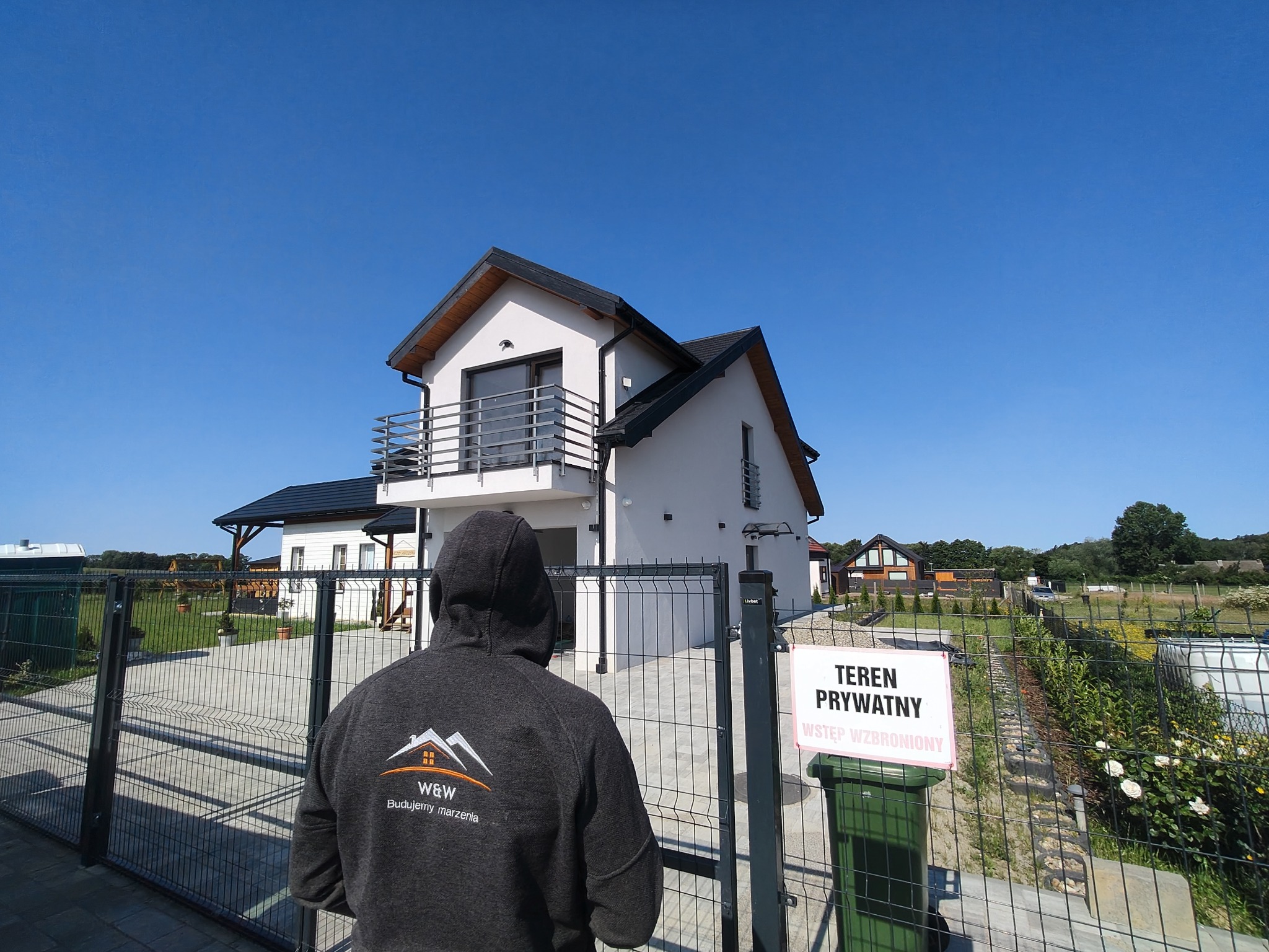 Nowoczesny dom jednorodzinny z balkonem, widziany zza ogrodzenia przez osobę w bluzie z logo firmy budowlanej na tle błękitnego nieba. Tabliczka 'Teren prywatny'.