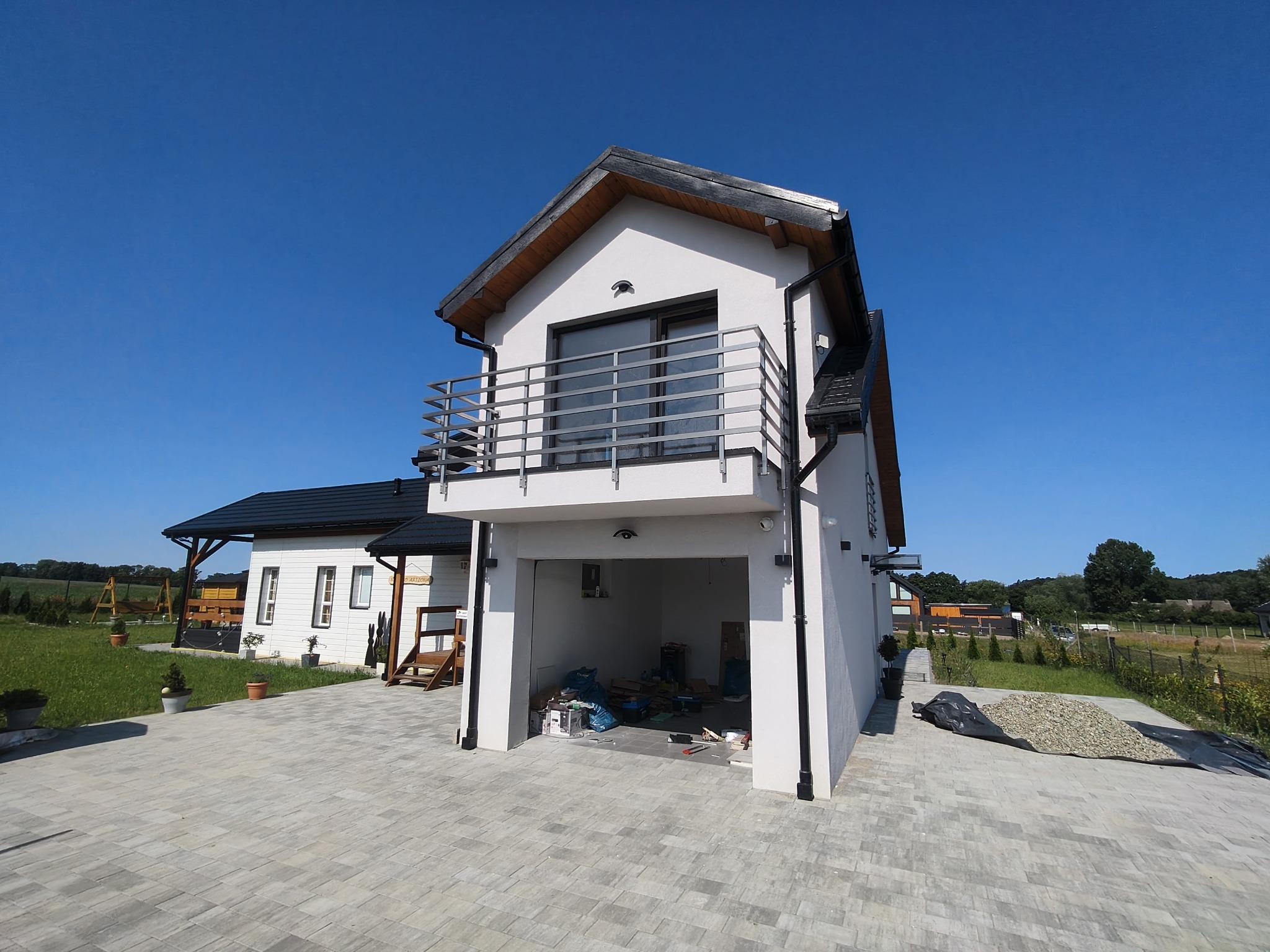 Dwukondygnacyjny dom z balkonem i garażem w stanie surowym otwartym, z widocznym bałaganem w garażu, na tle błękitnego nieba i zieleni. Ułożona kostka brukowa przed budynkiem.