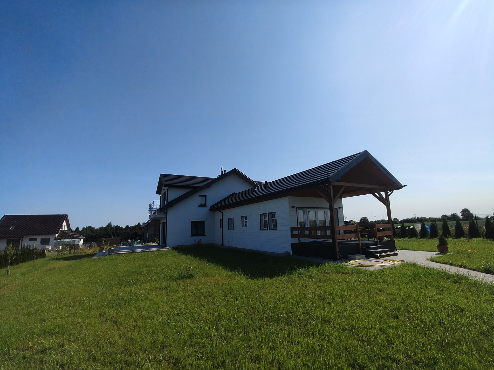 Nowoczesny dom z białymi ścianami i ciemnym dachem, zadaszony taras, zadbany trawnik, błękitne niebo. Widok z poziomu trawy, w tle drugi dom.