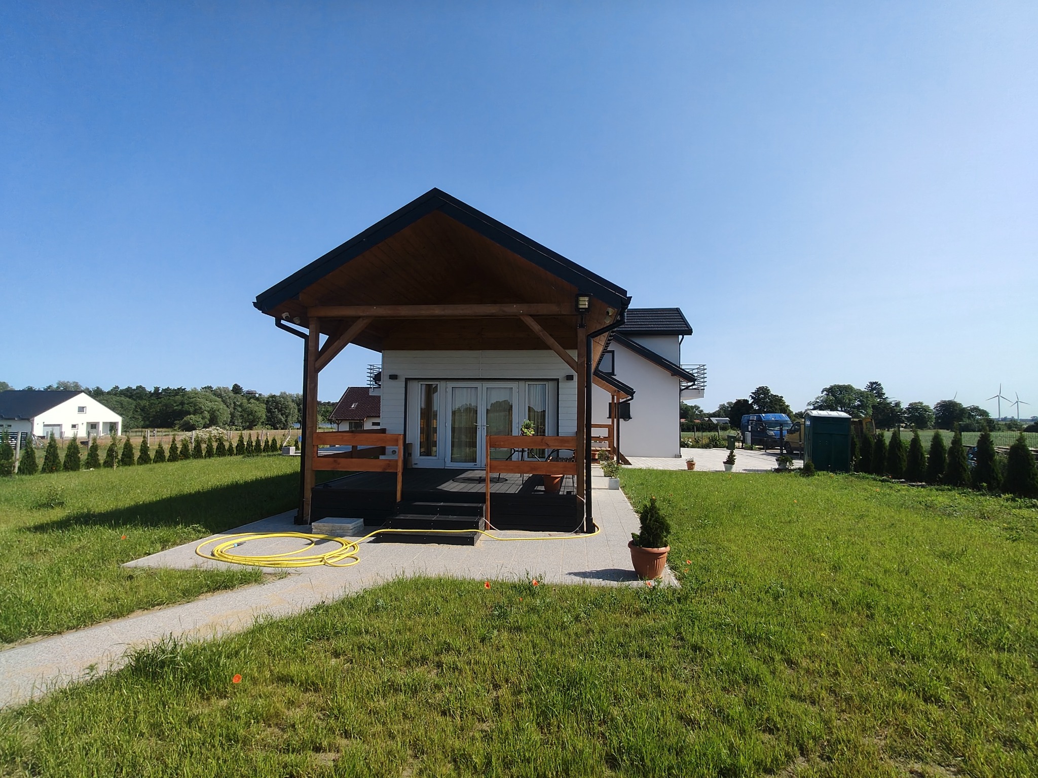 Nowoczesny dom z drewnianą werandą i zadbanym trawnikiem w słoneczny dzień. Żółty wąż ogrodowy leży na ścieżce. W tle widoczne inne domy i drzewa.