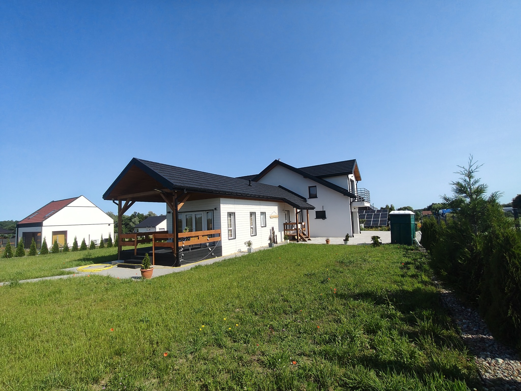 Nowoczesny dom z białymi ścianami i ciemnym dachem, z zadaszonym tarasem i zadbanym trawnikiem. Widoczne panele słoneczne w tle. Ujęcie w słoneczny dzień.