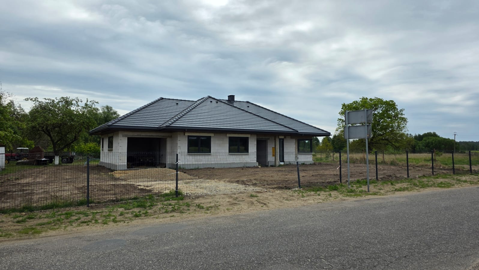 Niedokończony dom jednorodzinny z szarego bloczka, czarnym dachem i oknami, otoczony siatką. Widoczne przygotowane podłoże i elementy krajobrazu.