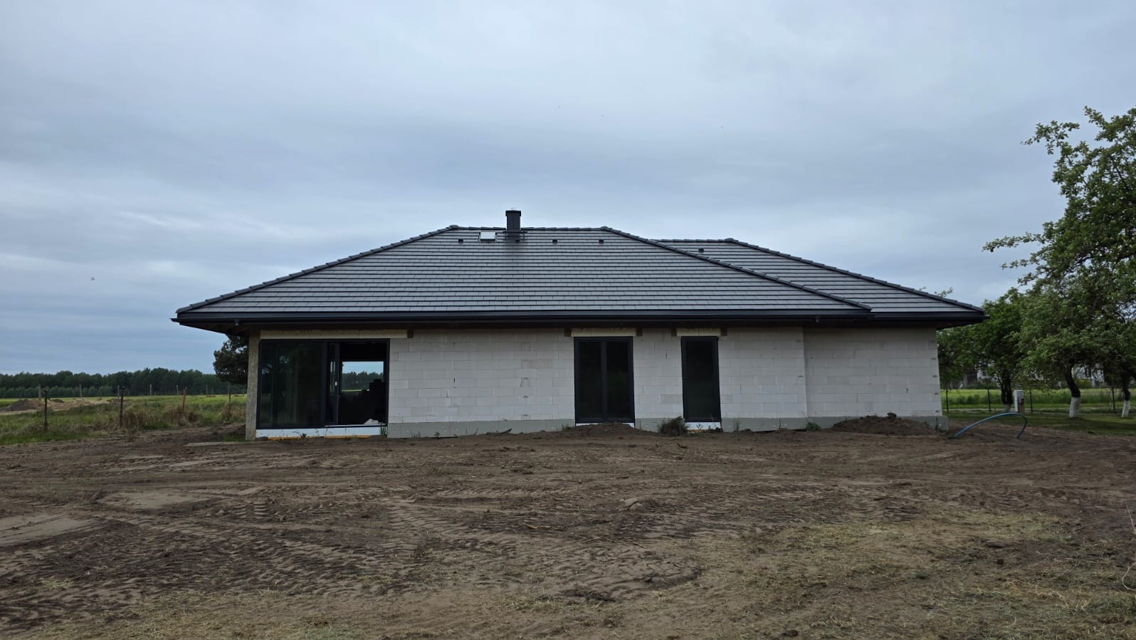 Niedokończony dom jednorodzinny z szarego bloczka, z ciemnoszarą dachówką, czarnymi oknami i drzwiami, na rozkopanym terenie. Widok z boku, pochmurne niebo.