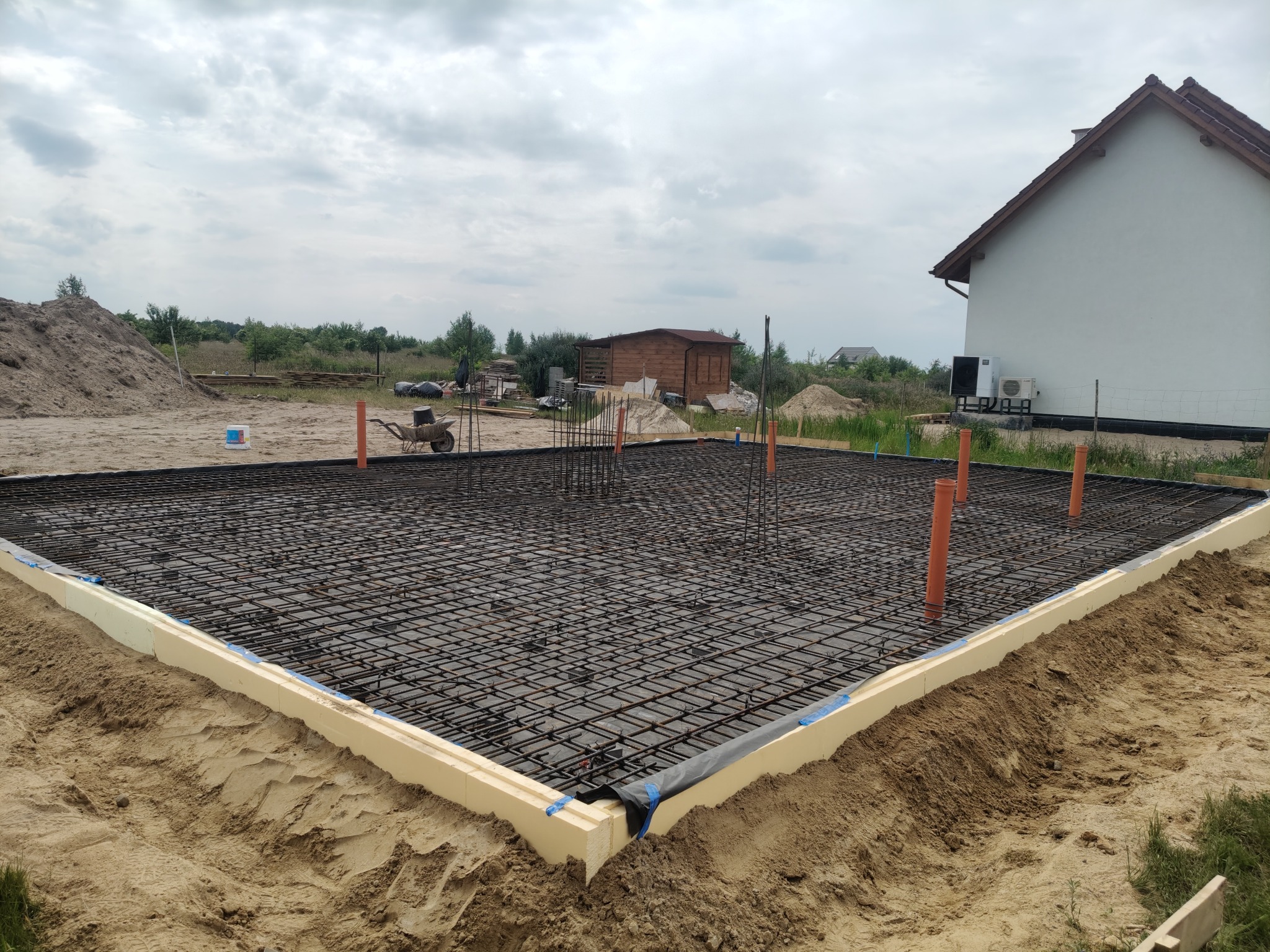 Zbrojenie fundamentu domu jednorodzinnego z widoczną izolacją termiczną i wystającymi rurami instalacyjnymi. W tle budynek w stanie surowym otwartym.