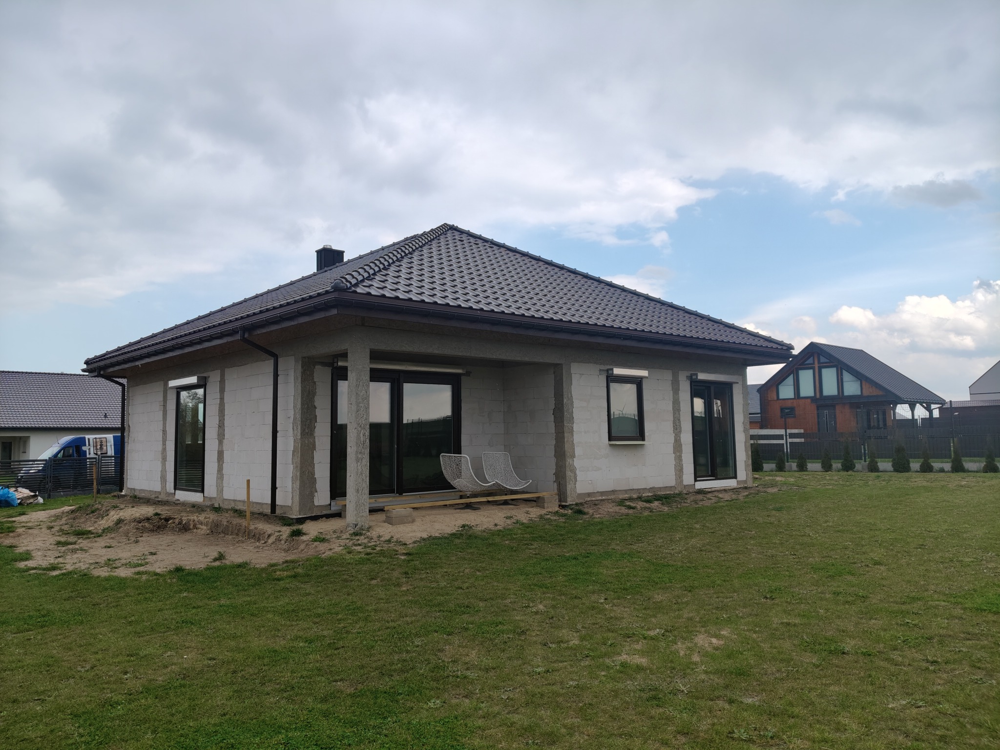 Surowy dom jednorodzinny z szarej cegły, z ciemnym dachem i dużymi oknami, w otoczeniu zielonego trawnika i innych domów w tle, w trakcie budowy.