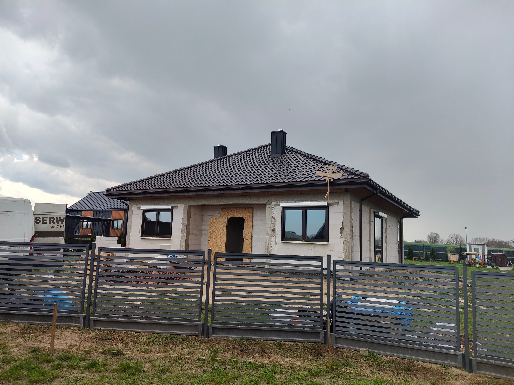 Surowy dom jednorodzinny z czarną dachówką, widoczny przez metalowe ogrodzenie. Częściowo otynkowane ściany, okna z ciemnymi ramami, zabezpieczone wejście.