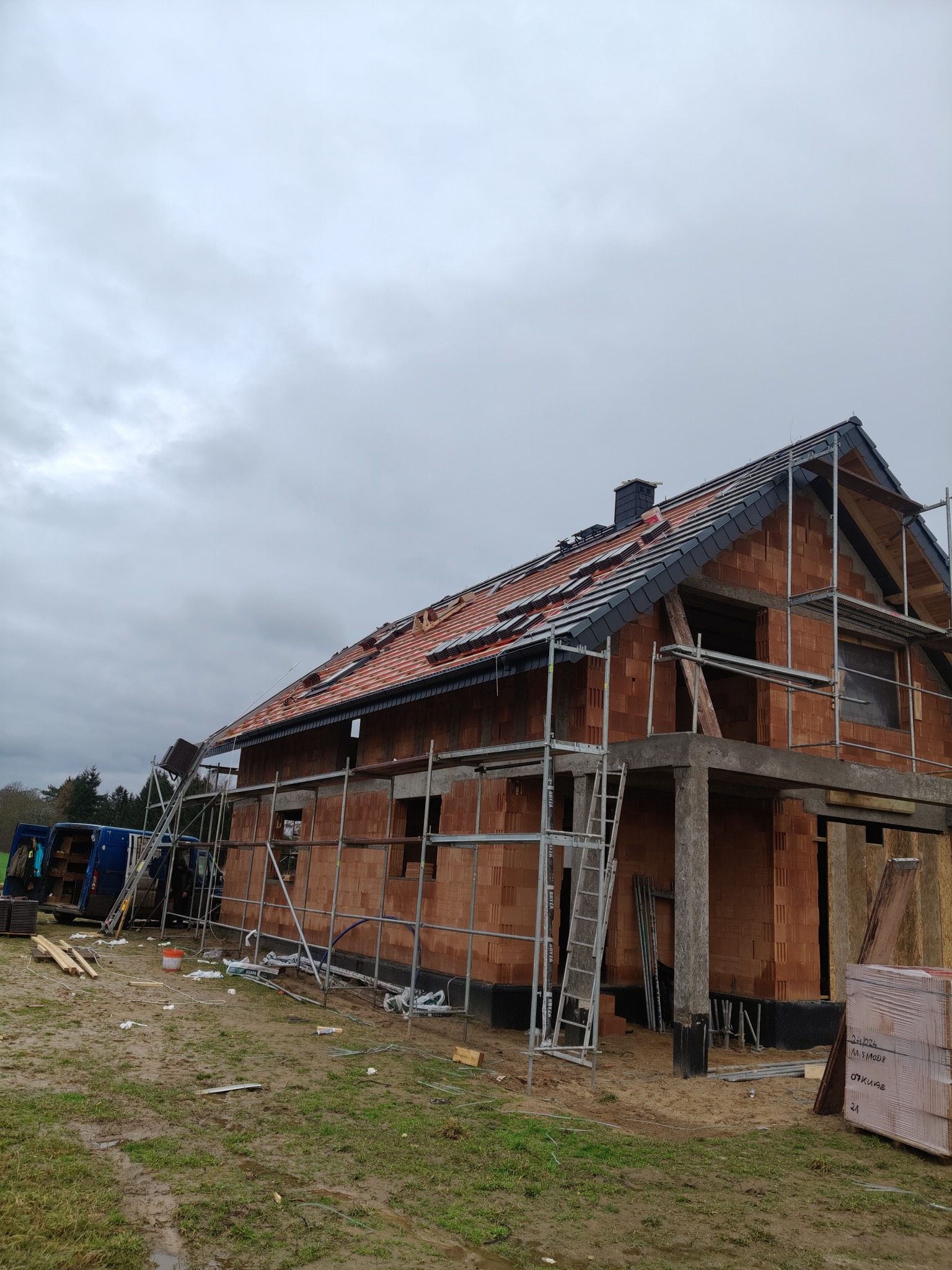 Budowa domu z czerwonej cegły w trakcie krycia dachu, widoczne rusztowania i częściowo ułożona dachówka na tle pochmurnego nieba. Etap prac dekarskich.