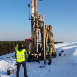 Wiercenie w zimowych warunkach: Dwóch pracowników w odblaskowych kamizelkach obsługuje wiertnicę na ośnieżonym terenie, widoczne rury wiertnicze i las w tle.
