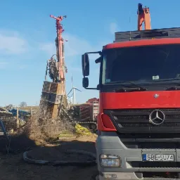 Czerwona wiertnica na placu budowy z widocznym ciężkim sprzętem i wiatrakami w tle, z bliska widoczny fragment ciężarówki Mercedes.