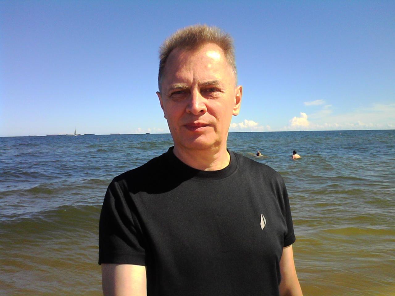 Mężczyzna w czarnym t-shircie na tle morza z pływającymi osobami i statkami w oddali, w słoneczny dzień.