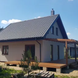 Nowoczesny dom z szarą dachówką i drewnianym tarasem, widok z boku na tle drzew i błękitnego nieba.