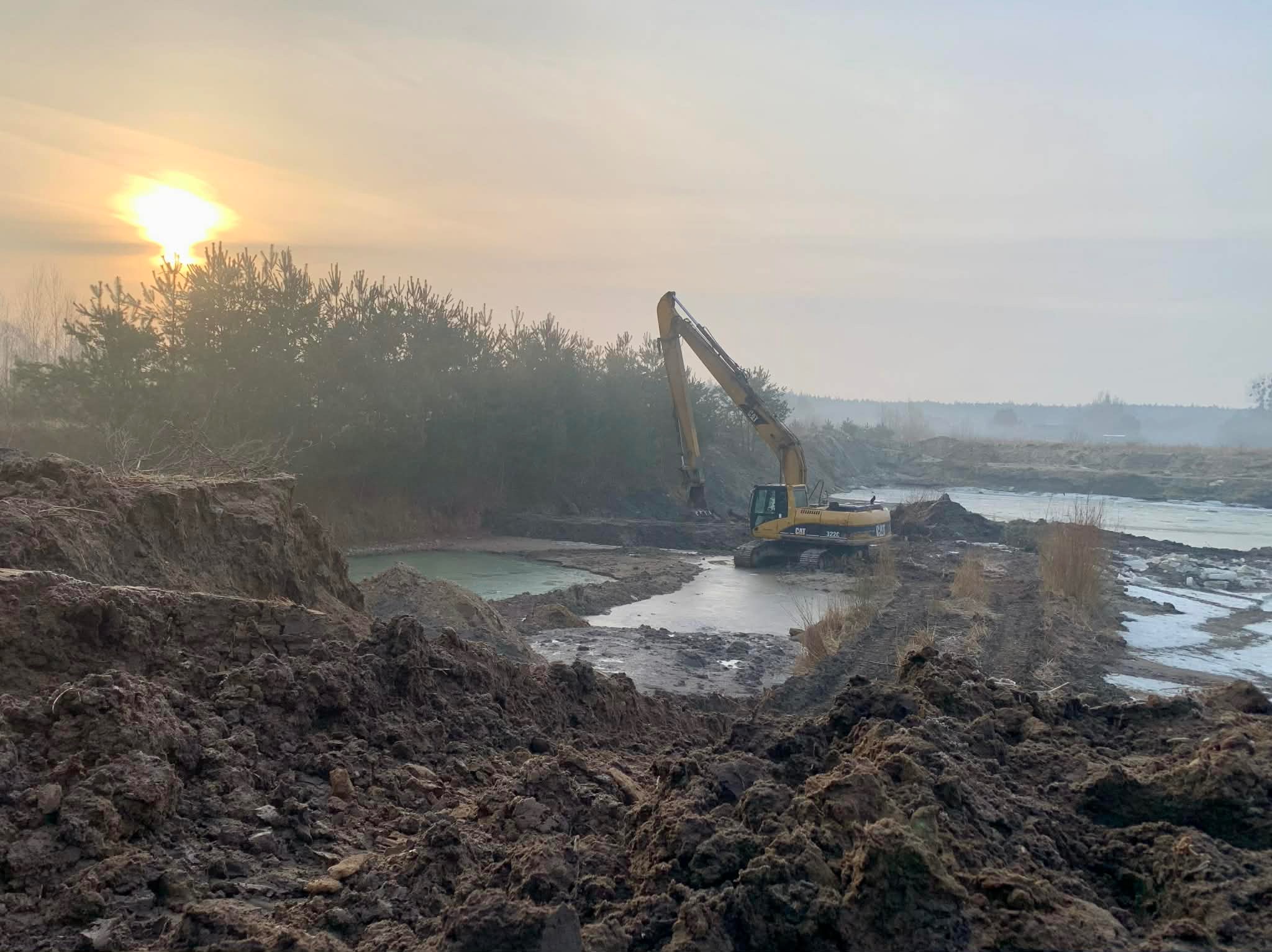 Żółta koparka CAT 322C przy wykopie stawu w mglisty poranek. Świecące słońce w tle, ziemia i woda. Widok na wykopany teren z bliska.