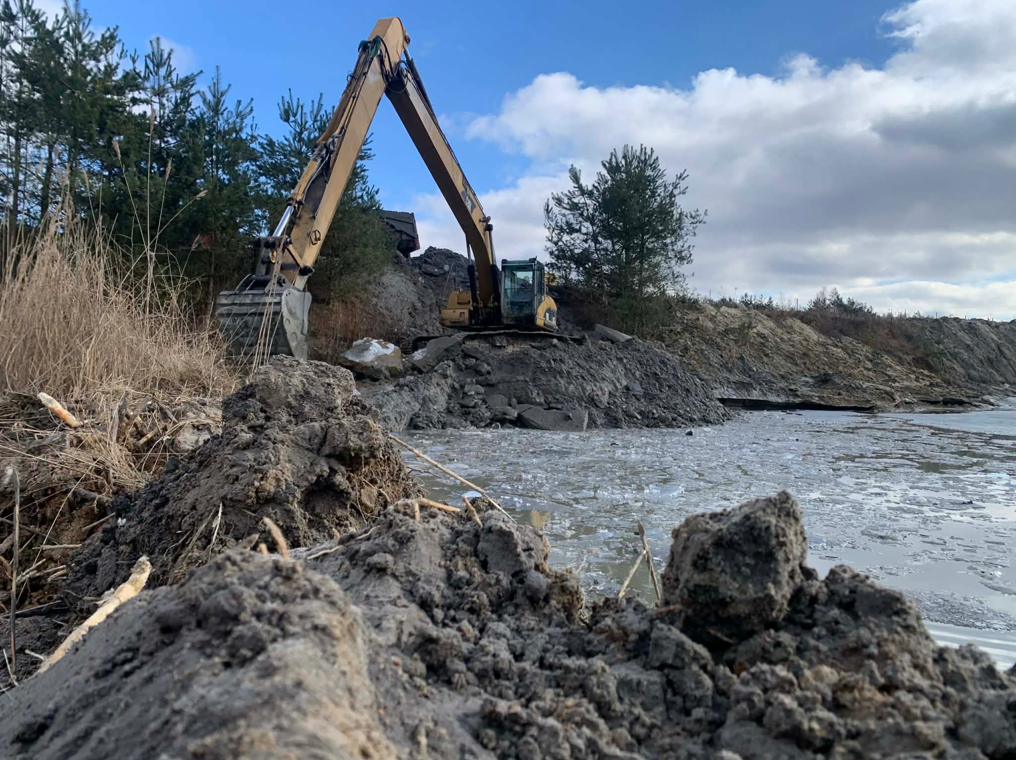 Żółta koparka na brzegu zbiornika wodnego, w tle drzewa i pochmurne niebo. Wykopy ziemne, prace ziemne przy zbiorniku wodnym.