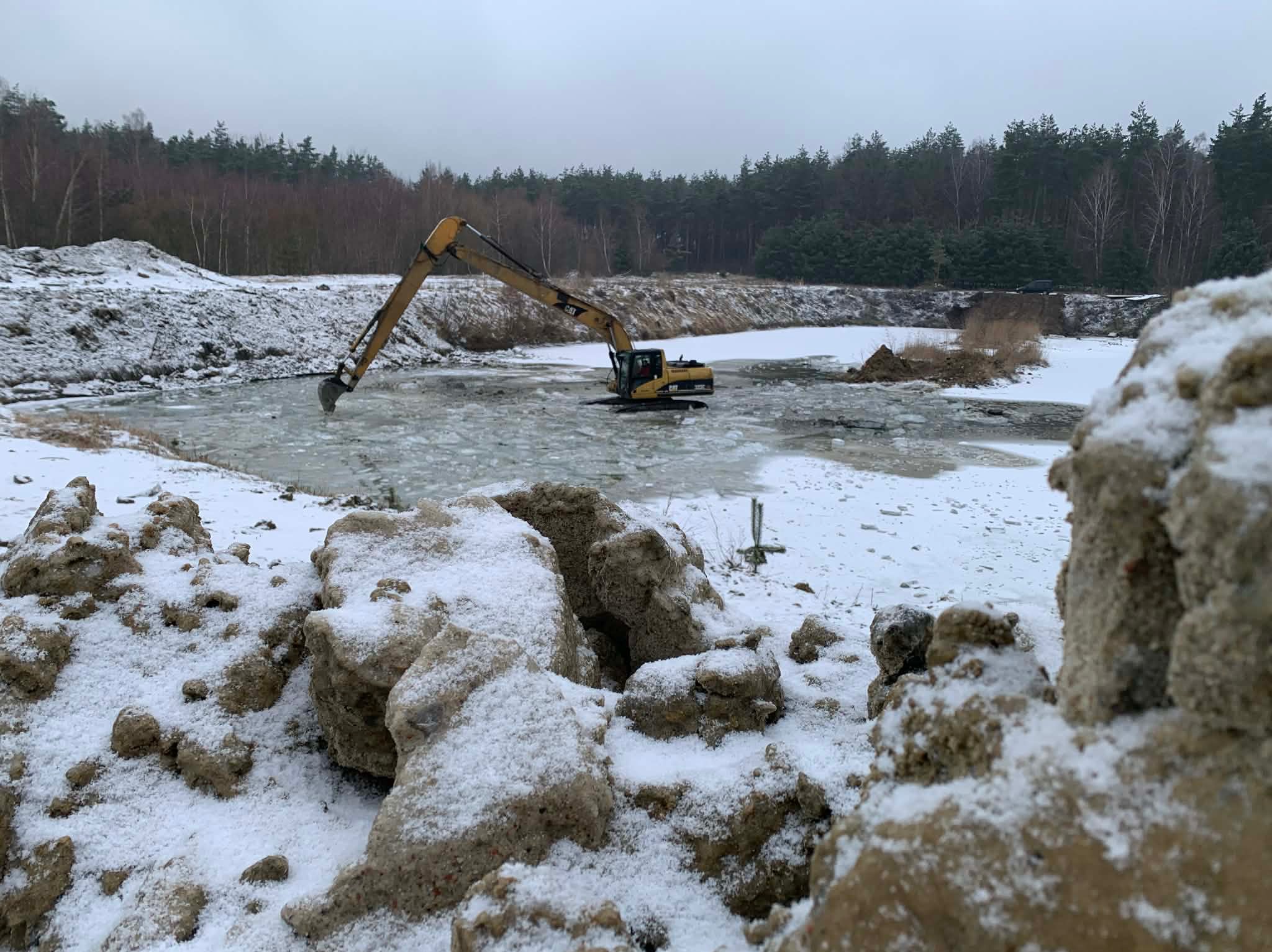 Żółta koparka CAT w zamarzniętym stawie, otoczona śniegiem i lasem. Ujęcie z bliska na wykopaną ziemię w pierwszym planie, podkreślające zimowy krajobraz.