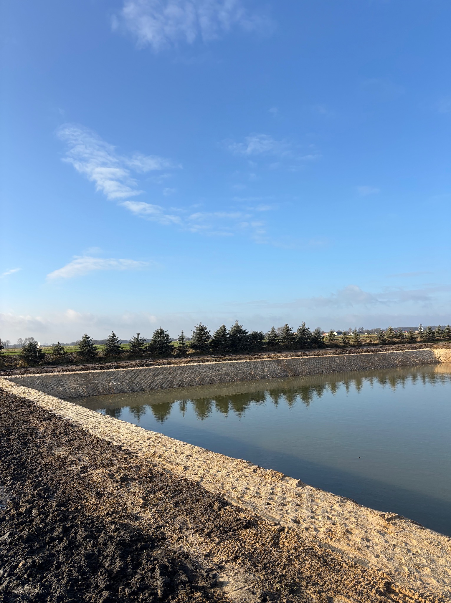 Wykopany staw z umocnionymi brzegami, obsadzony rzędem małych drzew iglastych, odbijających się w tafli wody pod błękitnym niebem z chmurami.
