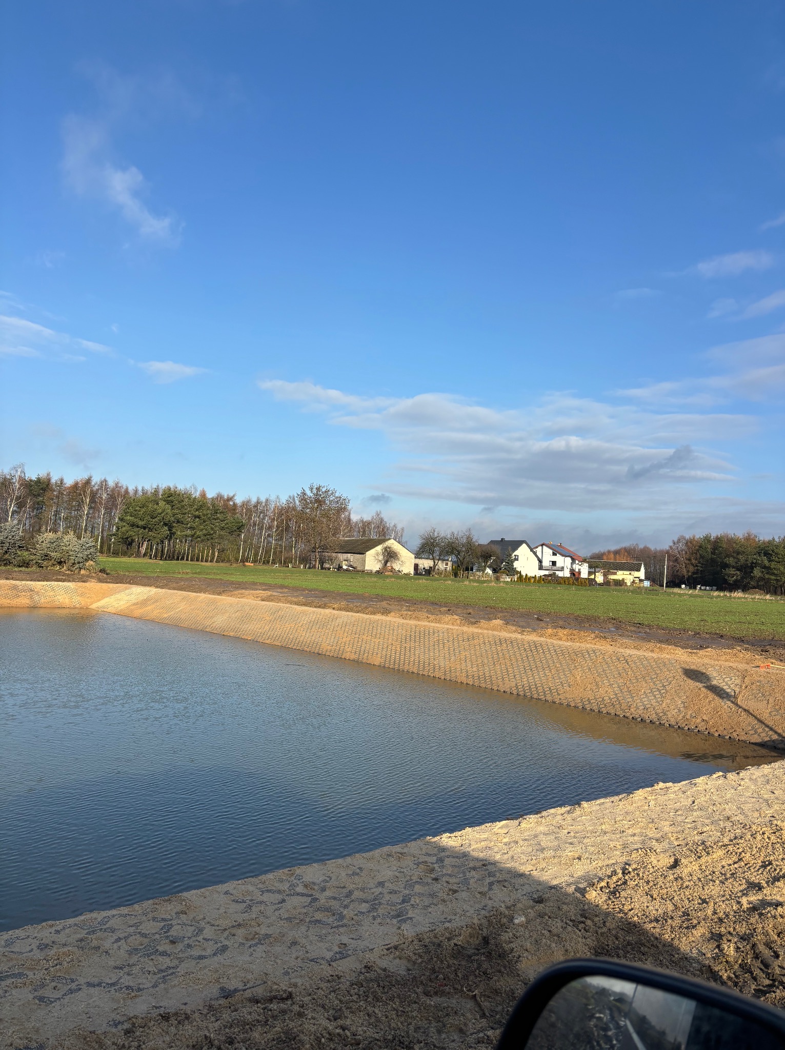Nowo wykopany staw z zabezpieczonym brzegiem, widok z perspektywy ziemi, w tle zabudowania i drzewa, błękitne niebo z chmurami.