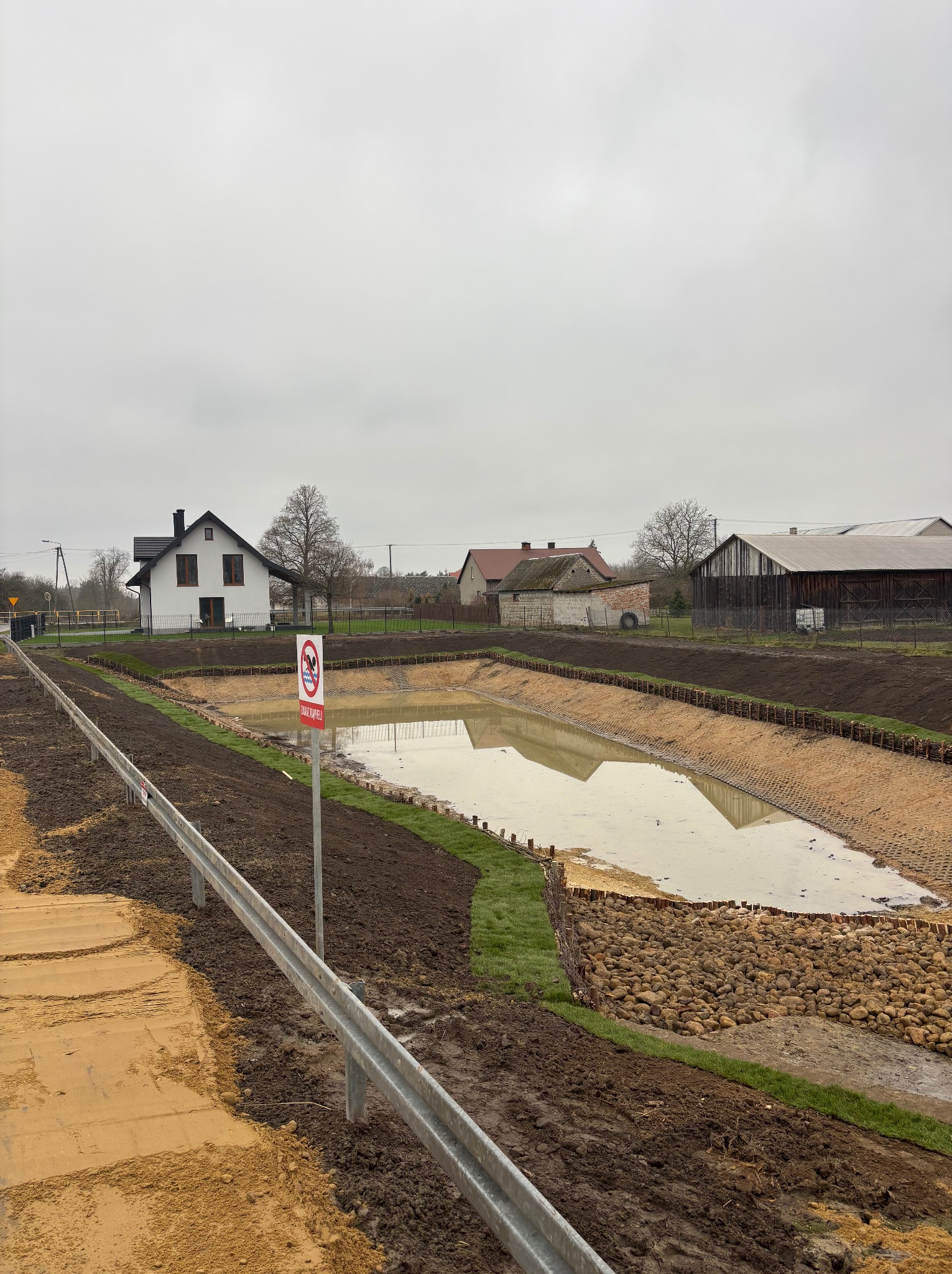Wykopany staw z zabezpieczonymi brzegami, częściowo wypełniony wodą. W tle zabudowania wiejskie i znak zakazu kąpieli. Ujęcie z boku, widoczne elementy infrastruktury.