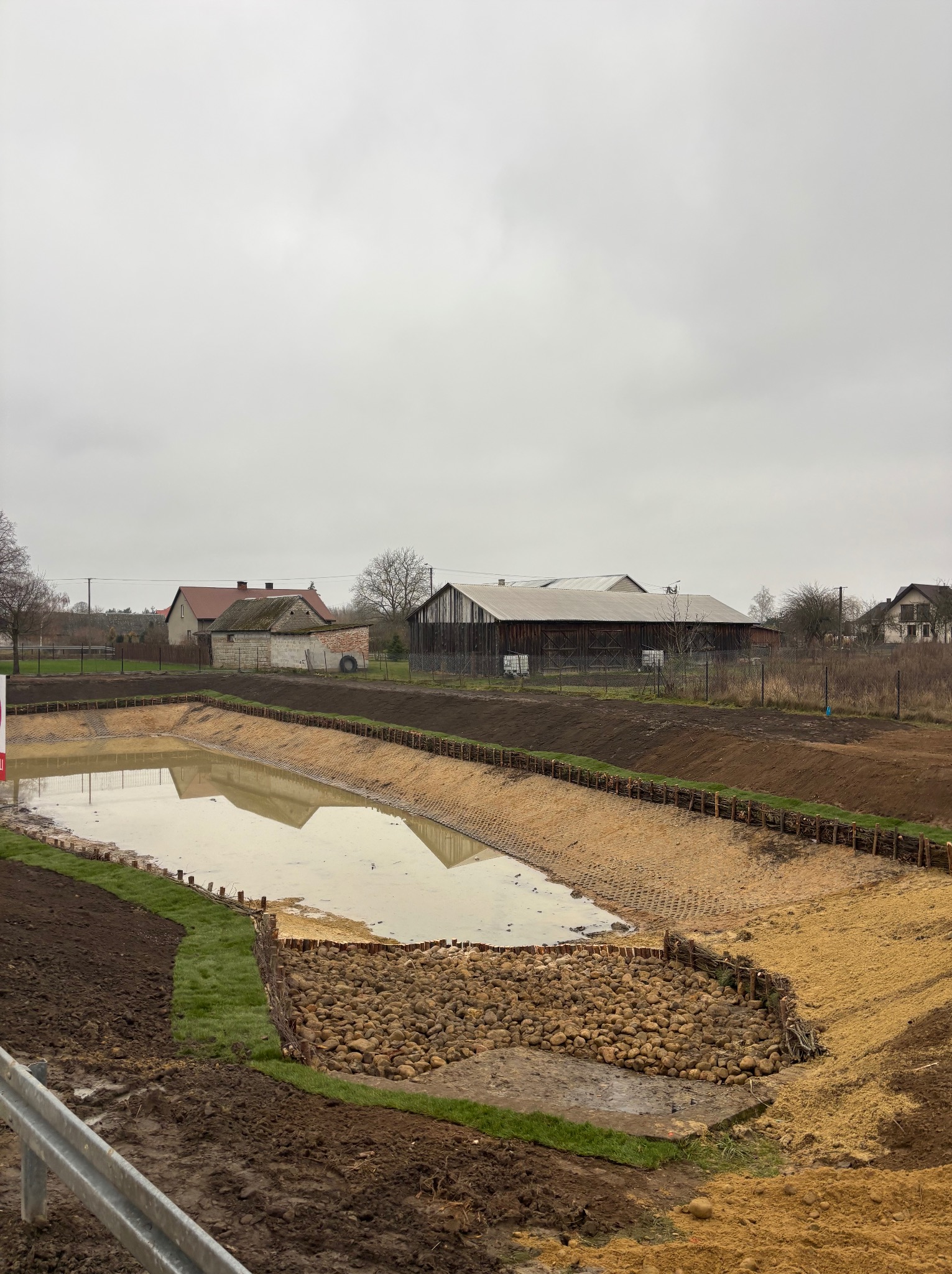 Nowo wykopany staw z zabezpieczonymi brzegami, wyłożonymi kamieniami i siatką. W tle zabudowania wiejskie pod pochmurnym niebem.