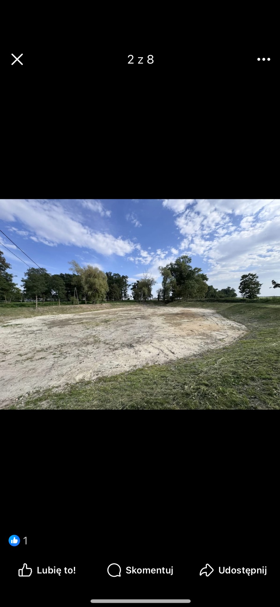 Wykarczowany teren pod budowę stawu, widoczne ślady po maszynach, w tle drzewa i błękitne niebo z chmurami. Teren przygotowany do dalszych prac ziemnych.
