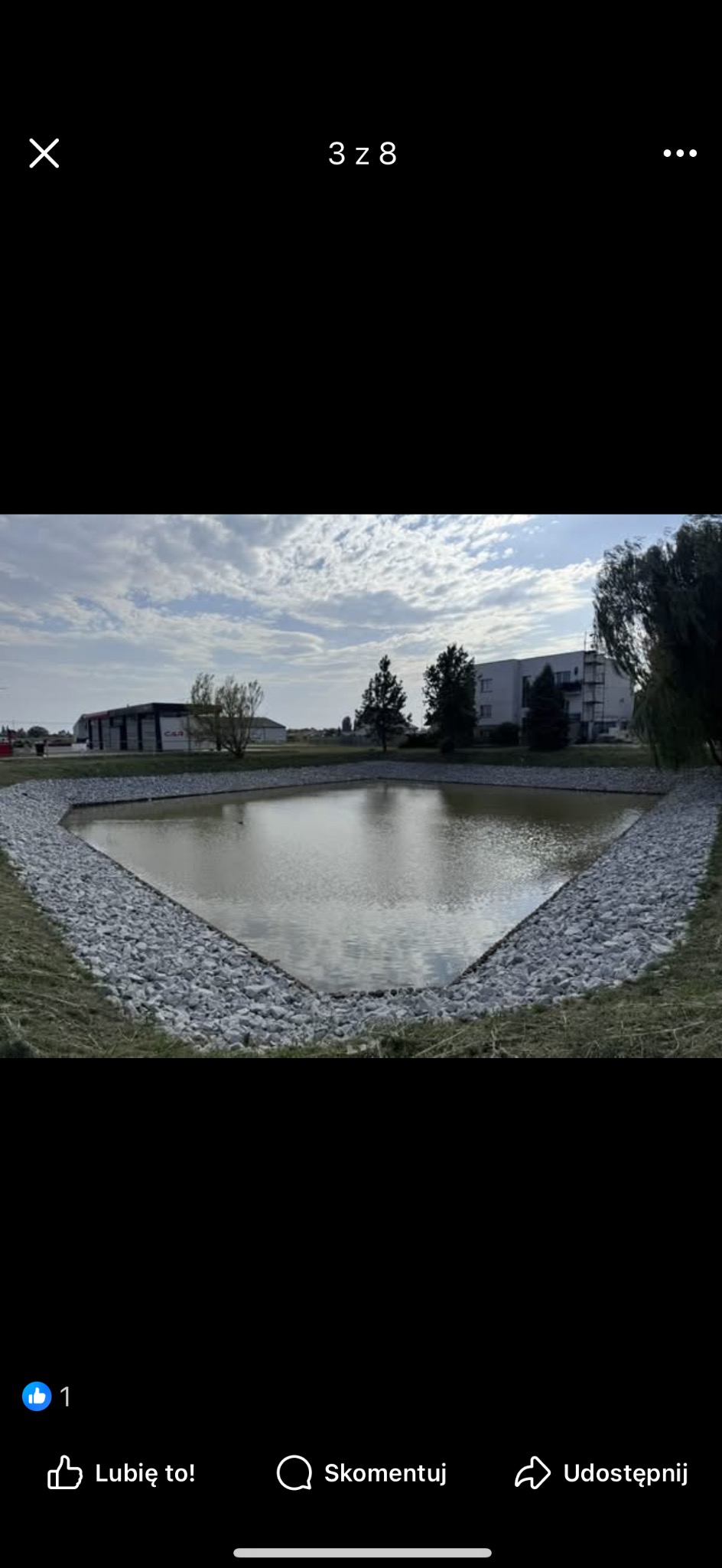 Nowo wykopany staw o trójkątnym kształcie, wyłożony kamieniami, otoczony zielenią. W tle budynki i błękitne niebo z chmurami.