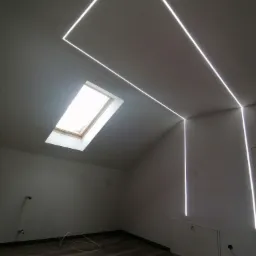 Poddasze z oknem dachowym i nowoczesnym oświetleniem LED w kształcie prostokąta biegnącym po suficie i ścianach, widoczne przewody i rękawice robocze na podłodze.
