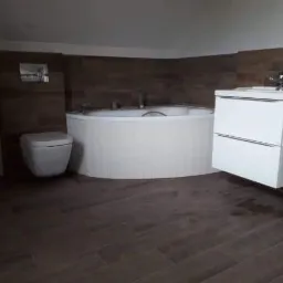 Nowoczesna łazienka z okrągłą wanną, podwieszaną toaletą i szafką z umywalką na tle drewnopodobnych płytek ściennych i podłogowych.