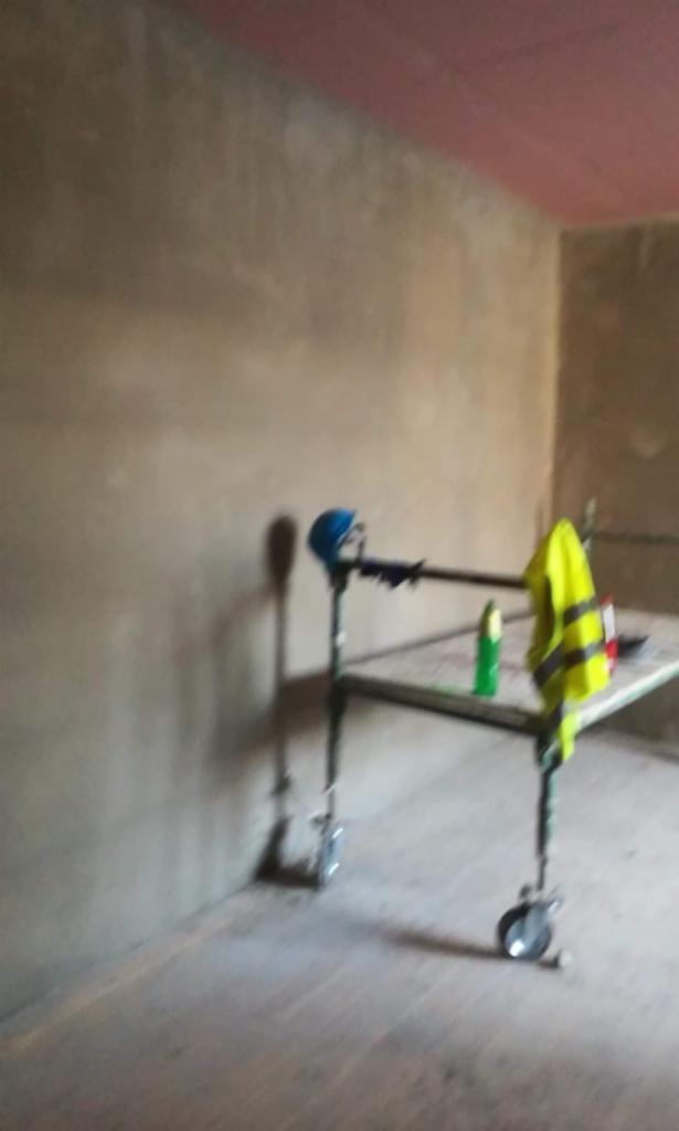 Wnętrze w trakcie prac wykończeniowych: surowa ściana, metalowe rusztowanie z kółkami, niebieski kask i żółta kamizelka odblaskowa.