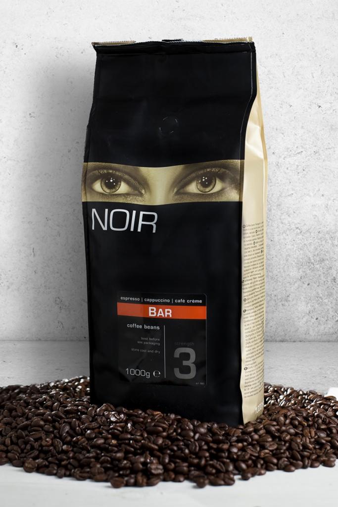 Czarna paczka kawy ziarnistej Noir Bar, o wadze 1000g, otoczona ciemnymi ziarnami kawy na neutralnym tle.